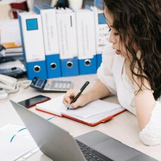 A person taking notes at a desk which is cluttered with office appliances