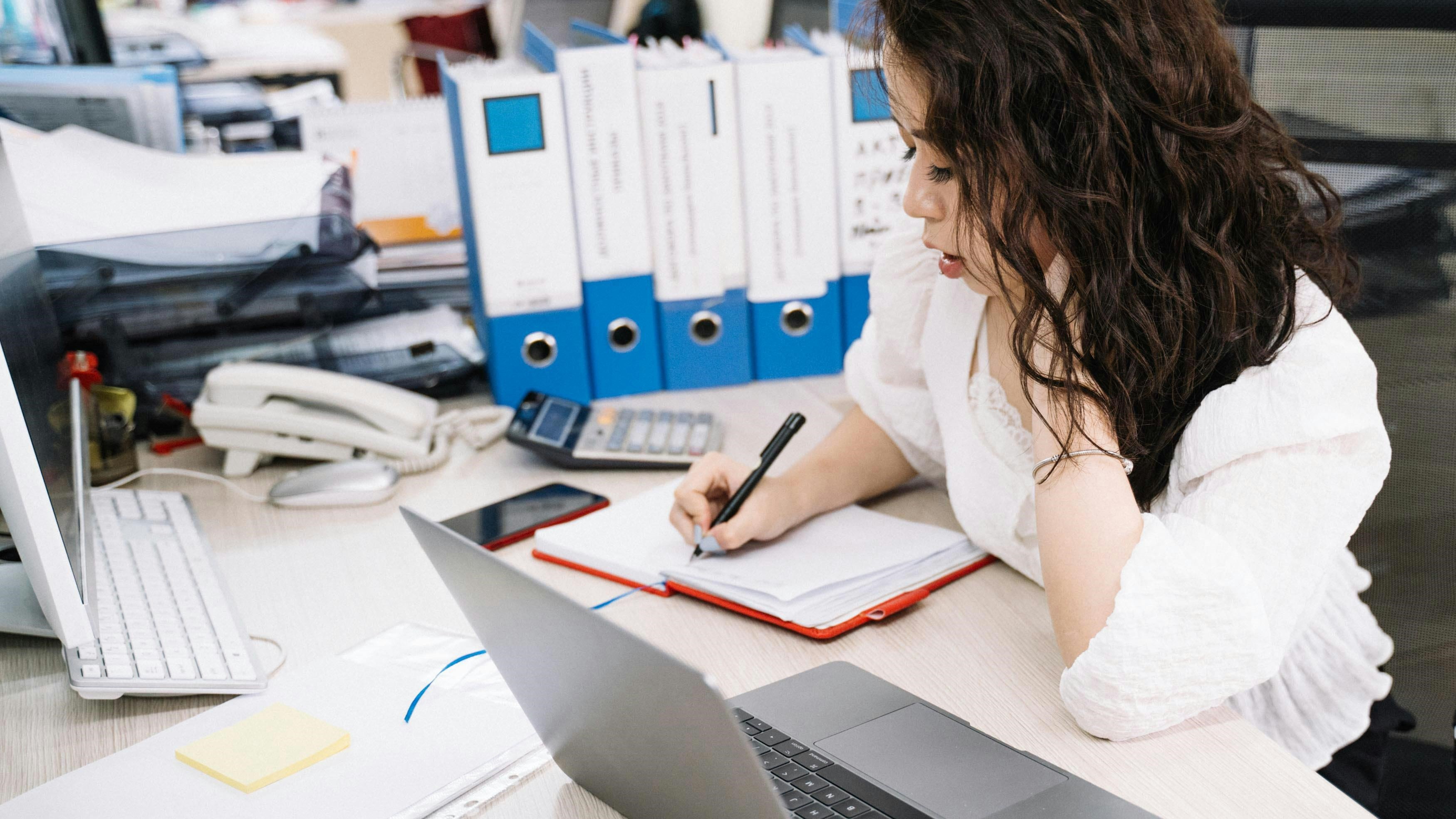 A young person is taking notes at an office desk which is cluttered with office supplies